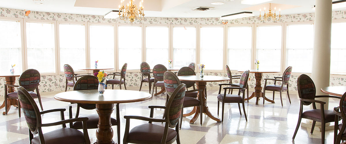 A bright and empty dining room with tables and chairs, illuminated by sunlight streaming through large windows.