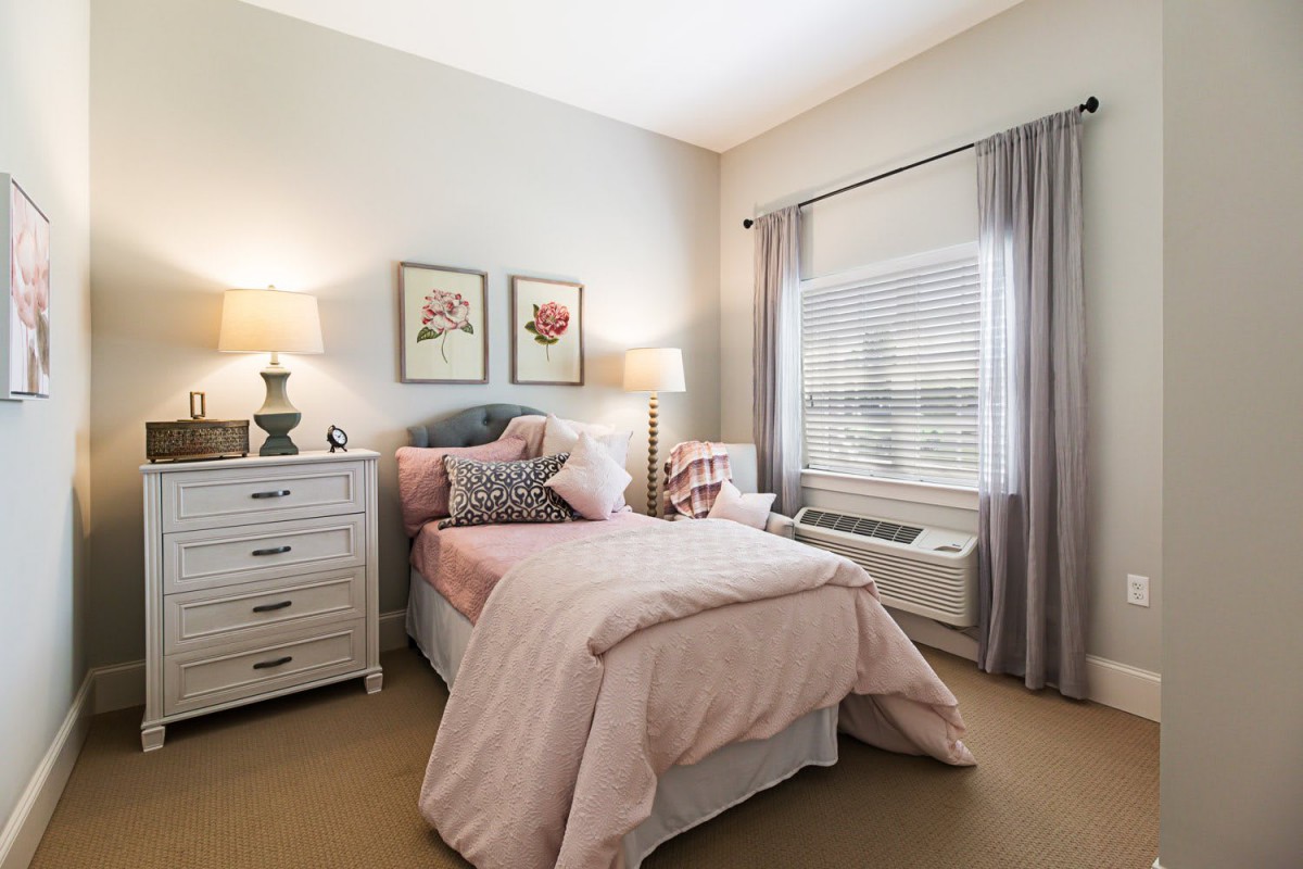 A cozy bedroom with soft pastel decor, featuring a bed with pink bedding, a dresser, and two lamps.