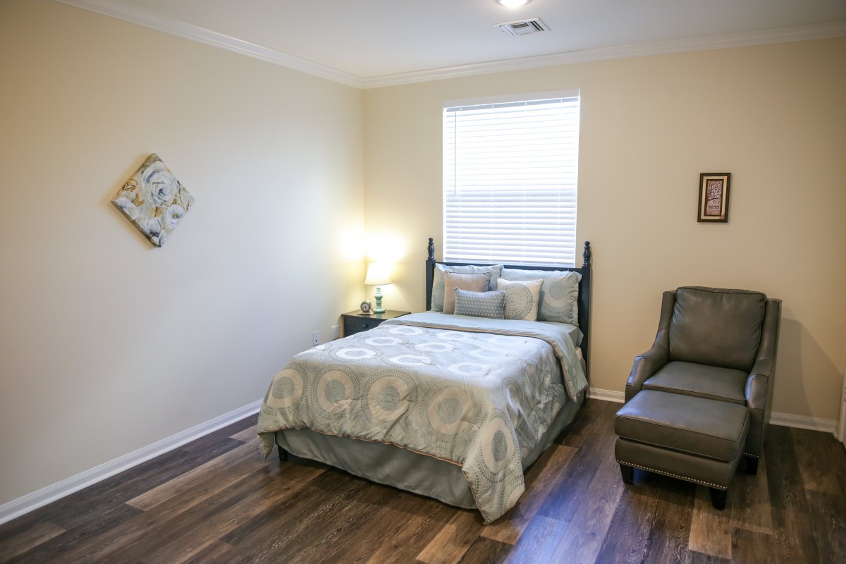 A cozy bedroom with a neatly made bed, an armchair, and a lamp beside the window.