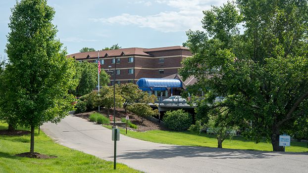 A building with a blue awning is surrounded by lush trees, with a road leading up to it and a flagpole visible in the foreground.