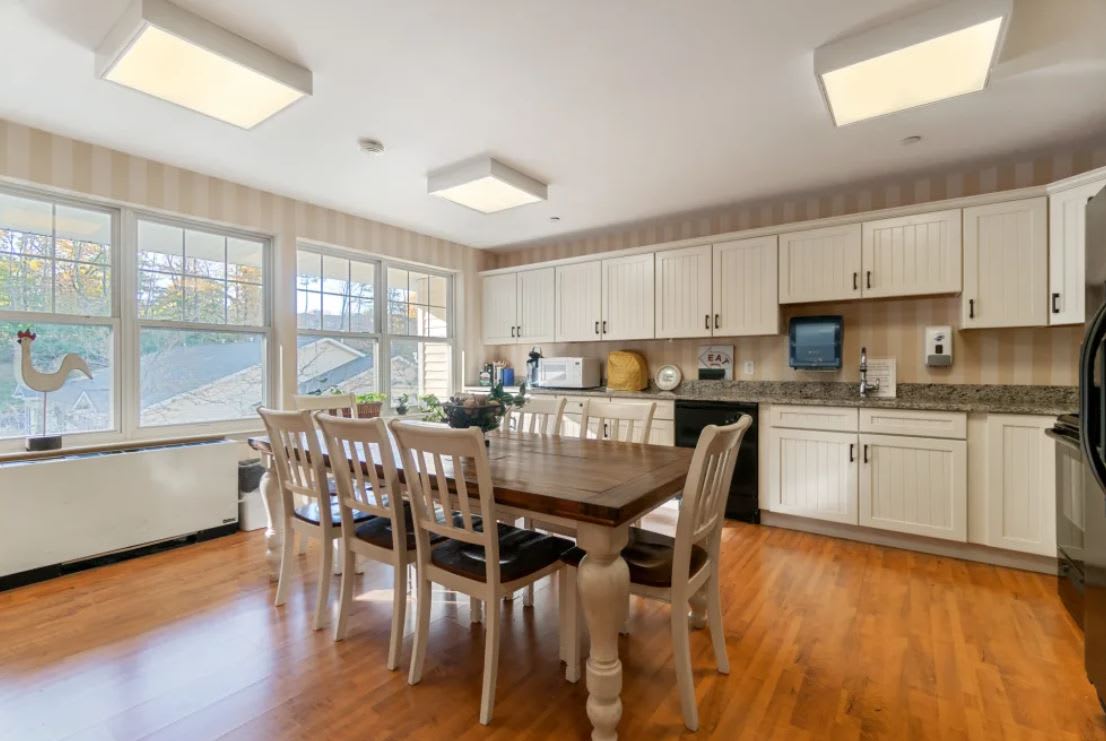 A bright kitchen and dining area with a wooden table and chairs, featuring white cabinets and ample natural light.