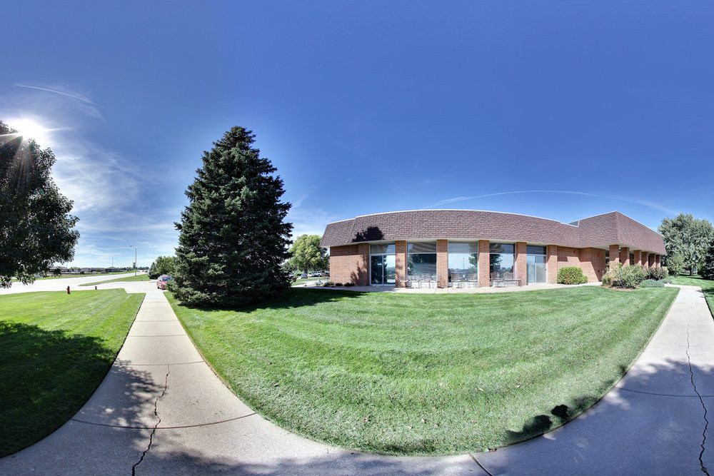 A brick building with a sloped roof adjacent to a large tree and a grassy area under a clear blue sky.