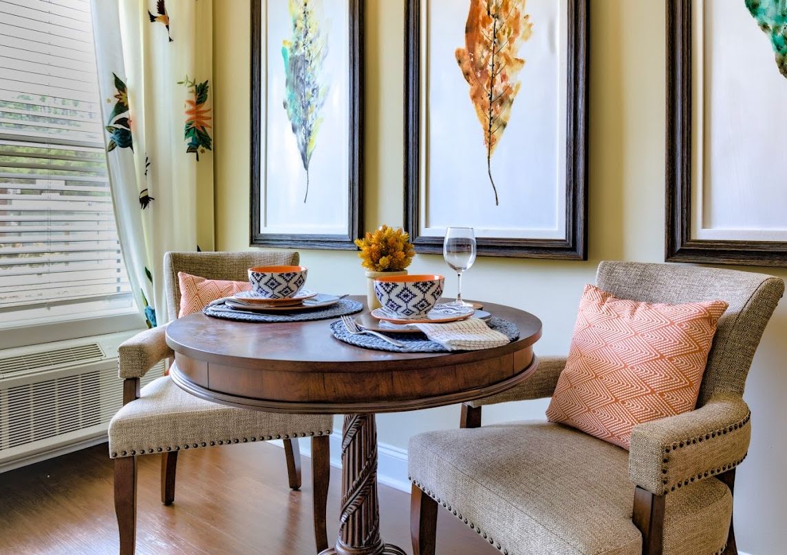A cozy dining area with a round wooden table set for two, complemented by framed leaf artwork and soft cushions.