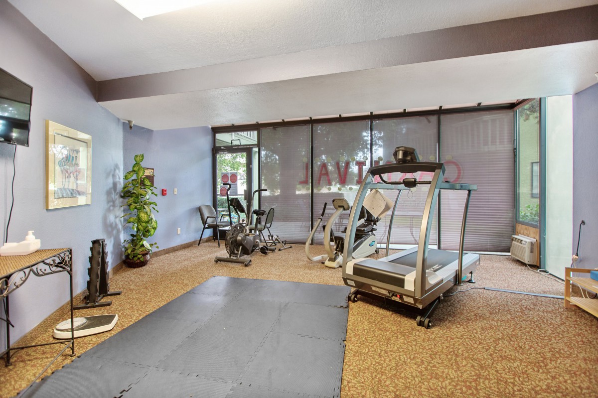 A well-lit gym room with exercise equipment including a treadmill, stationary bike, and an exercise mat.