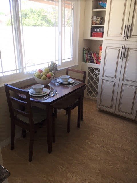 A cozy kitchen nook with a dining table set for two, featuring a fruit bowl and decorative shelves.