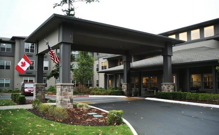 The image shows a building entrance with a covered driveway, featuring an American flag and surrounding landscaping.