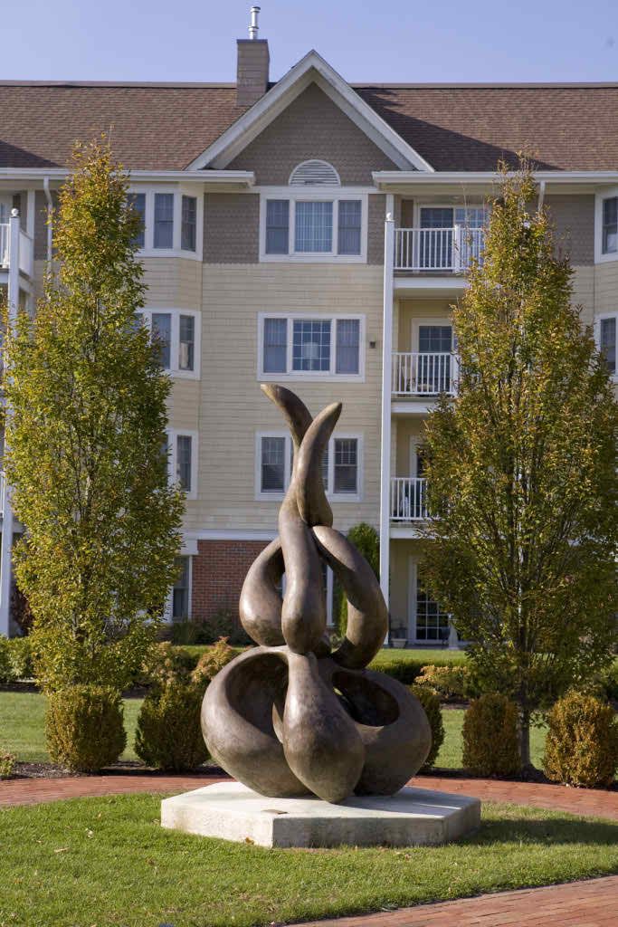 An abstract sculpture set in a landscaped area with trees and a residential building in the background.