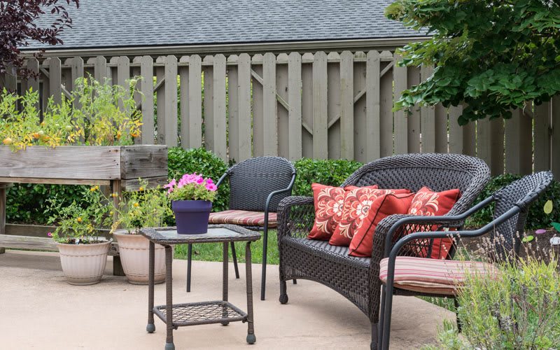 A cozy outdoor patio setup featuring wicker chairs with red cushions, a small table, and potted plants against a wooden fence backdrop.