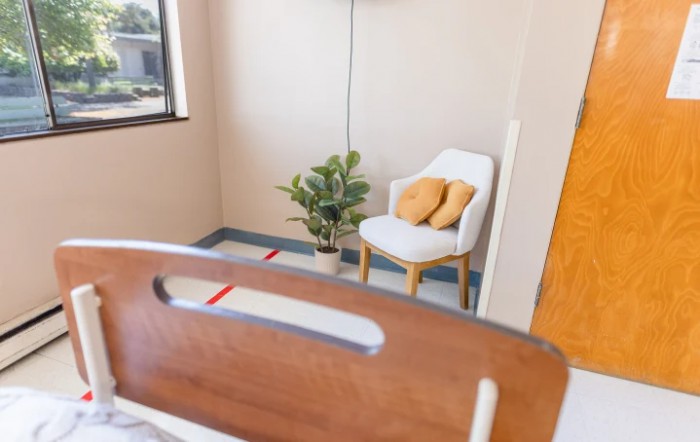 A cozy hospital room with a bed, a chair with pillows, and a potted plant by the window.