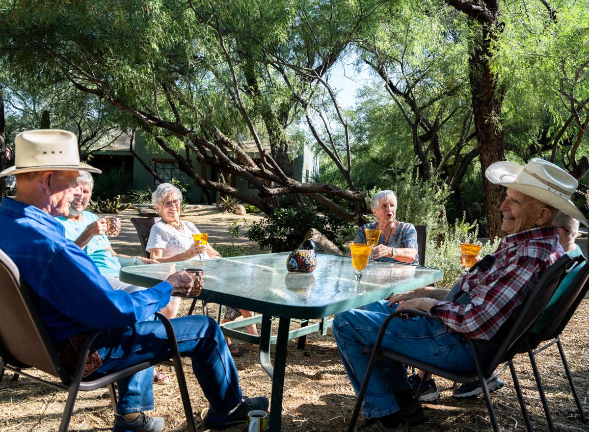 A group of elderly people enjoying drinks while sitting at a garden table surrounded by trees.