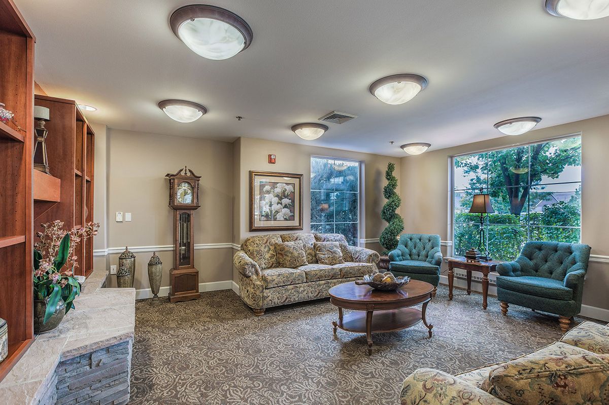 A cozy living room with traditional decor, featuring a sofa, coffee table, and a grandfather clock, enhanced by natural light from large windows.