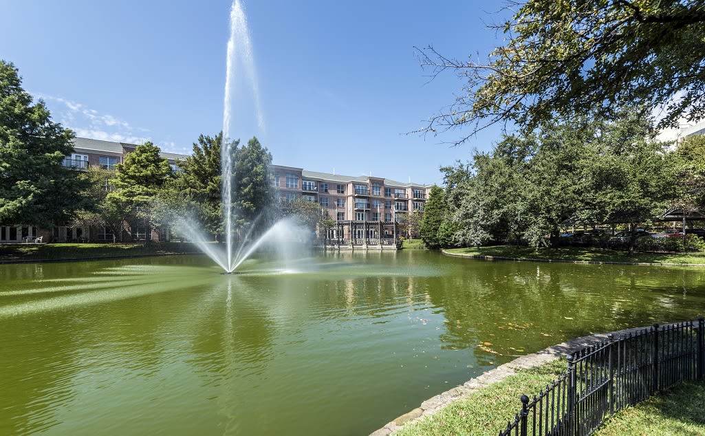 A scenic view of a fountain in a pond surrounded by trees and an apartment building.