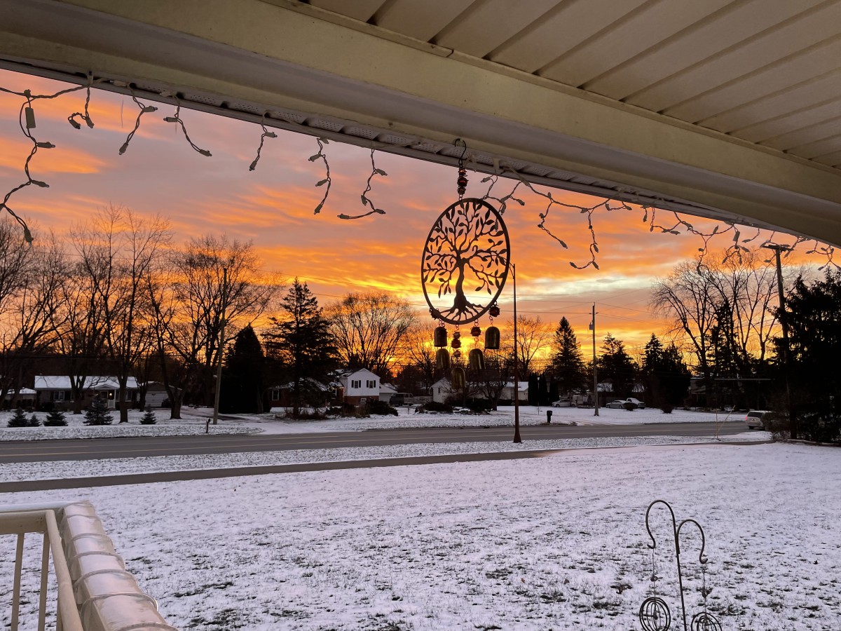 A beautiful winter sunset viewed from a snow-covered porch, with silhouettes of trees and houses in the distance.