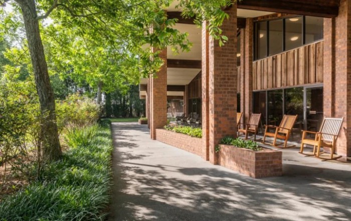 A sunny outdoor patio area with brick architecture and lush greenery along a sidewalk.