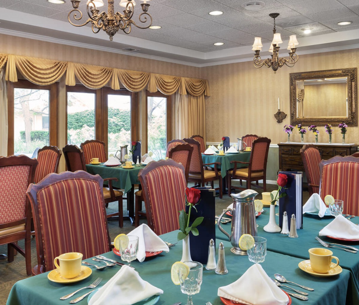 An elegantly decorated dining room with neatly arranged tables and chairs, featuring chandeliers and large windows.