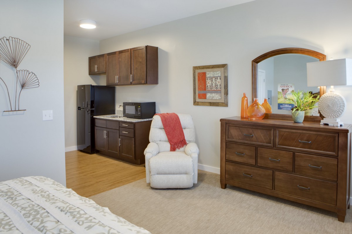 A cozy living area with a kitchenette, featuring a white recliner, dark wooden cabinets, and a dresser with a mirror.
