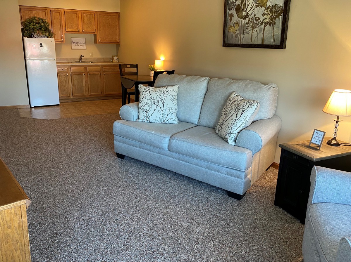 A cozy living room with a sofa and cushions, adjacent to a small kitchen area.