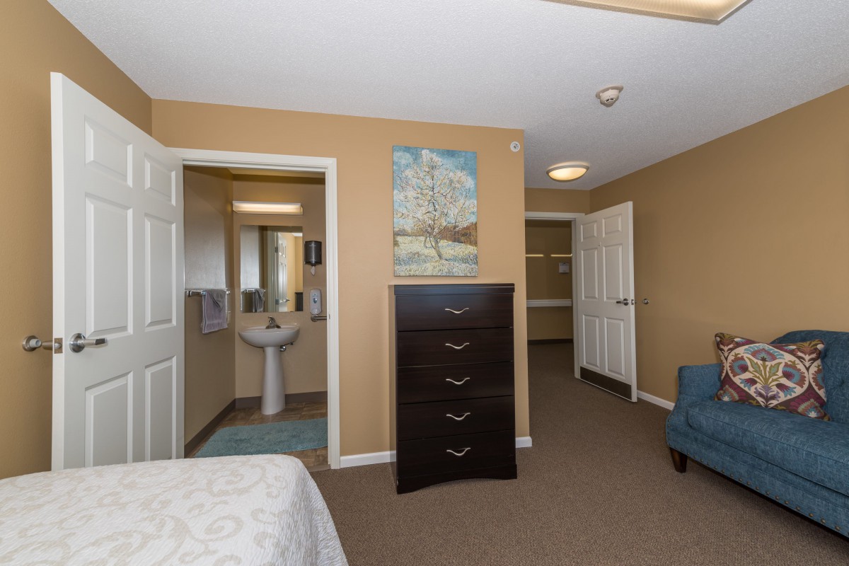 A cozy bedroom with a view into an adjacent bathroom, featuring a dresser and armchair.