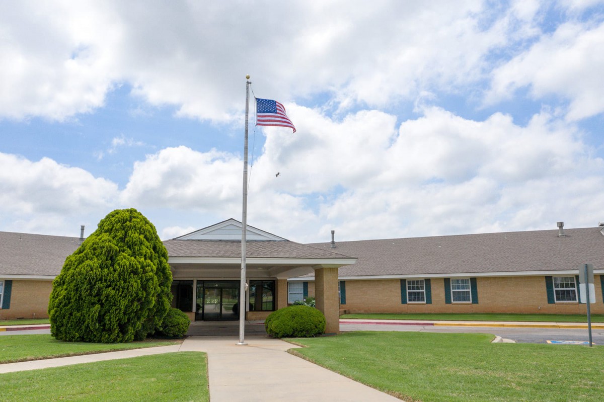 A school building with an American flag flying on a flagpole, surrounded by a lawn and pathway under a cloudy sky.