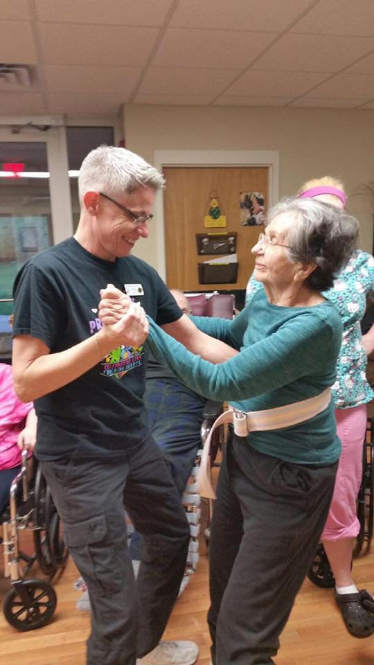 An elderly woman and a man dancing together, sharing a joyful moment in a community setting.