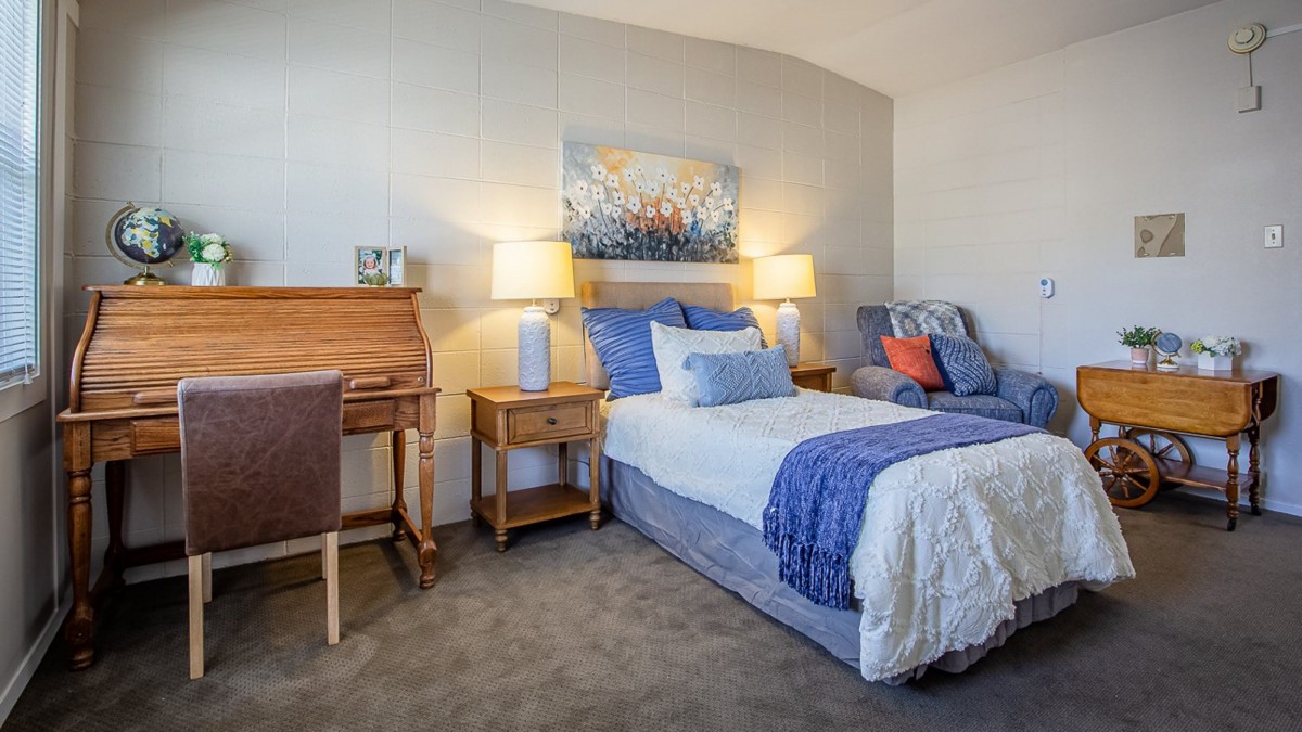 A cozy bedroom with a neatly made bed, decorated in blue and white tones, featuring a wooden desk and two table lamps.