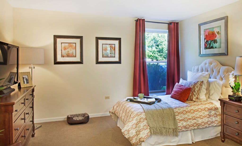 A cozy bedroom featuring a neatly made bed with a decorative blanket and pillows, two framed artworks on the wall, and a large window with red curtains.