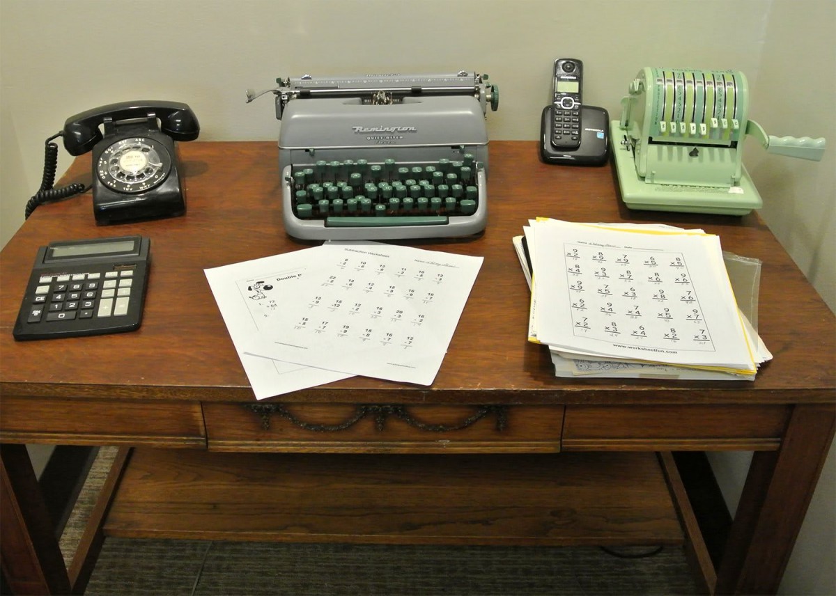 A vintage desk setup featuring a typewriter, rotary phone, calculator, and various papers.