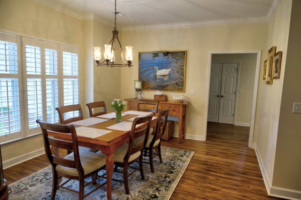 A cozy dining room features a wooden table with chairs, illuminated by an elegant chandelier. A painting decorates the wall above a sideboard, adding to the warm and inviting atmosphere.