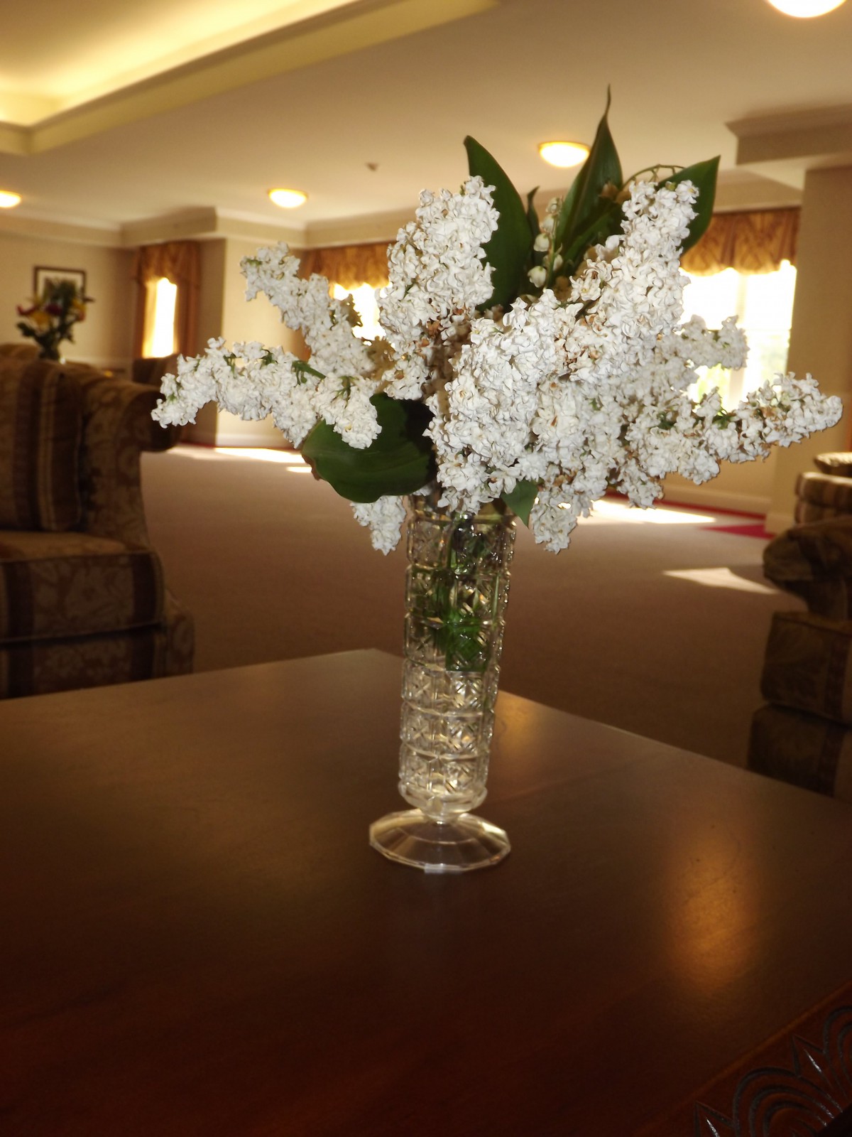 A vase of white lilac flowers on a table in a warmly lit interior.