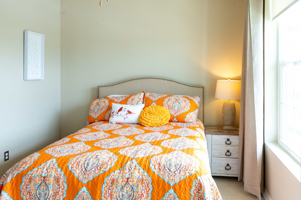 A cozy bedroom with an orange and white quilt on the bed, accompanied by decorative pillows and a bedside lamp.