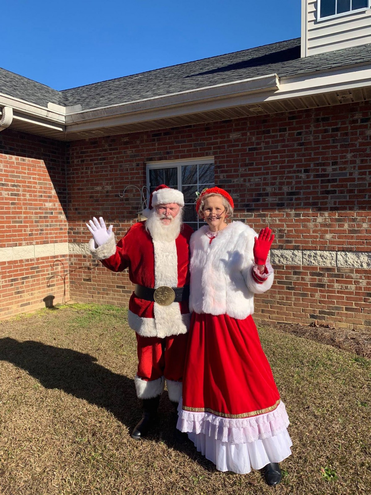 A cheerful Santa and Mrs. Claus wave outside a brick building on a sunny day.