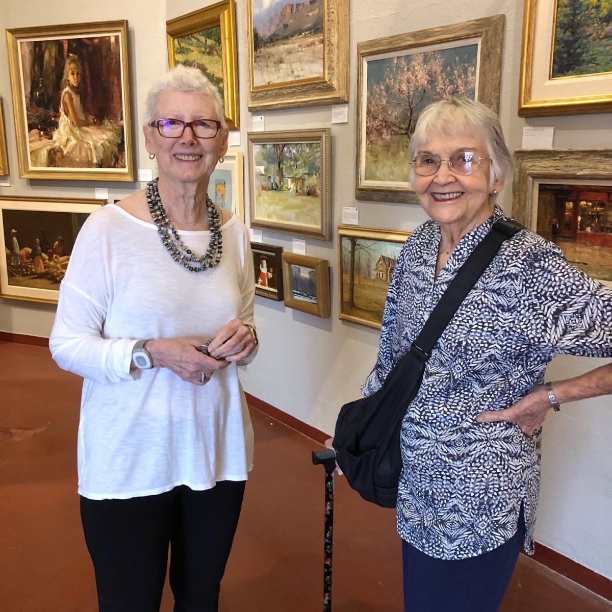 Two women standing in an art gallery surrounded by various paintings.