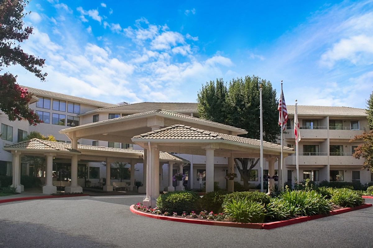 A large building with a covered entrance, surrounded by landscaping and flags, under a clear blue sky.