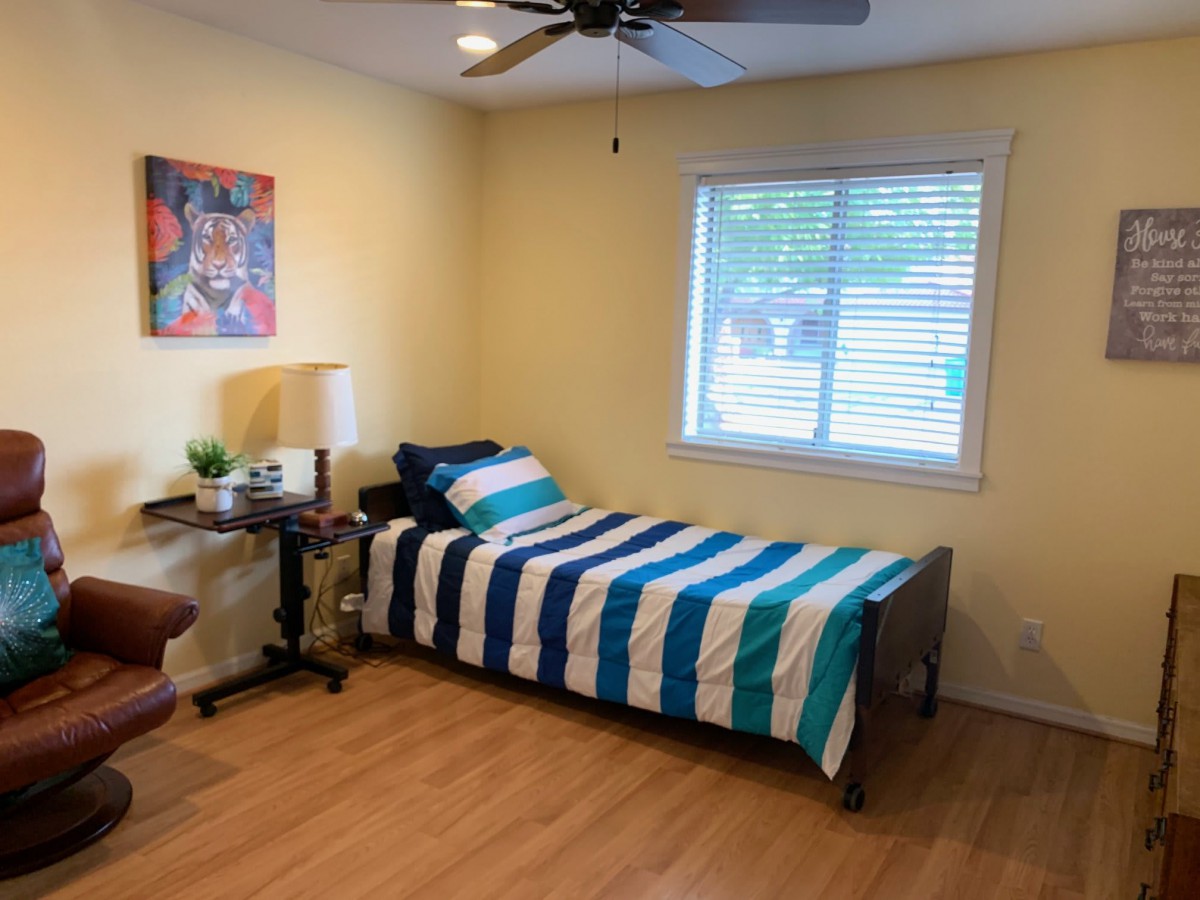 A cozy bedroom with a single bed featuring striped bedding, a desk and chair by the window, and a ceiling fan overhead.