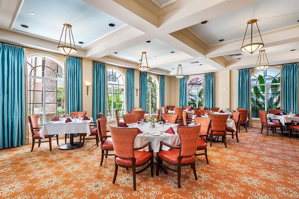 An elegant dining room with neatly arranged tables and chairs, featuring chandeliers and large windows with blue curtains.