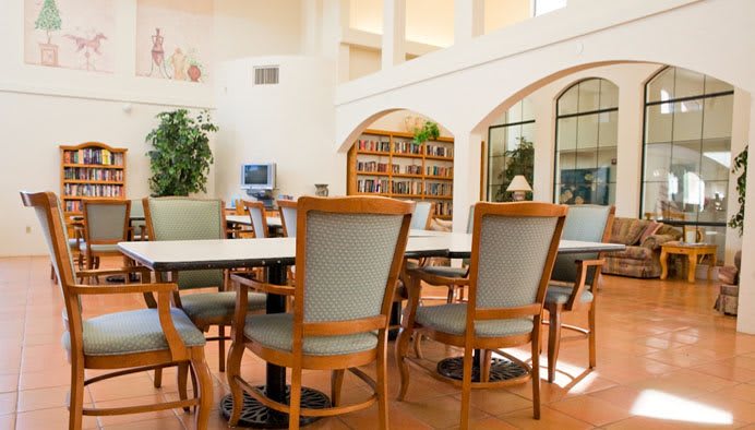A bright library space with tables and chairs, surrounded by bookshelves and plants.