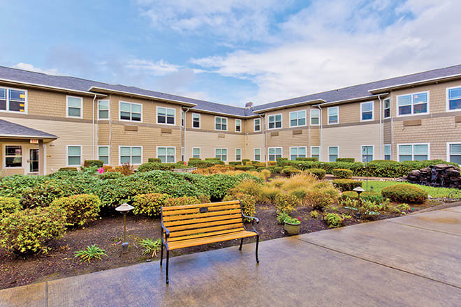A courtyard view of a two-story building with landscaped gardens and a bench under a partly cloudy sky.