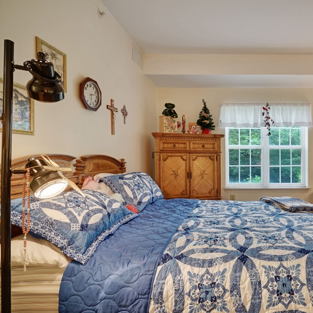 A cozy bedroom with a large bed featuring a blue patterned quilt, decorated walls, and a wooden dresser beside a window.