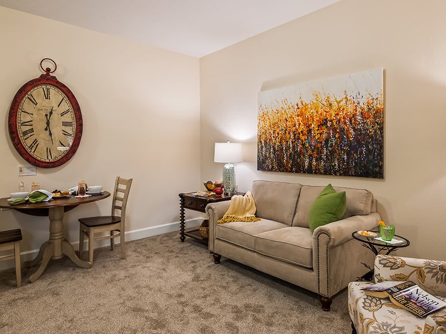 A cozy living room with a sofa, artwork on the wall, a large round wall clock, and a small dining area with table and chairs.