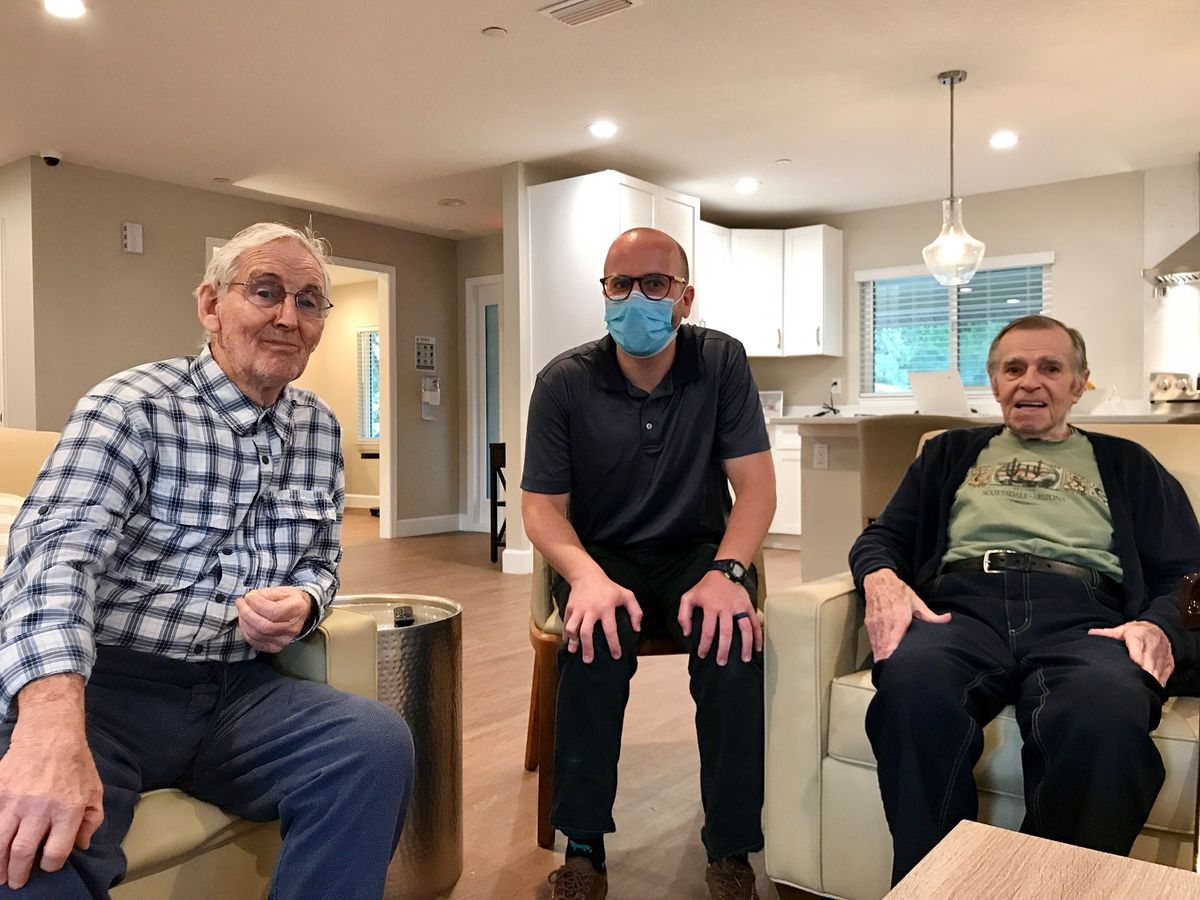 Three men sitting together in a living room setting, one wearing a mask.