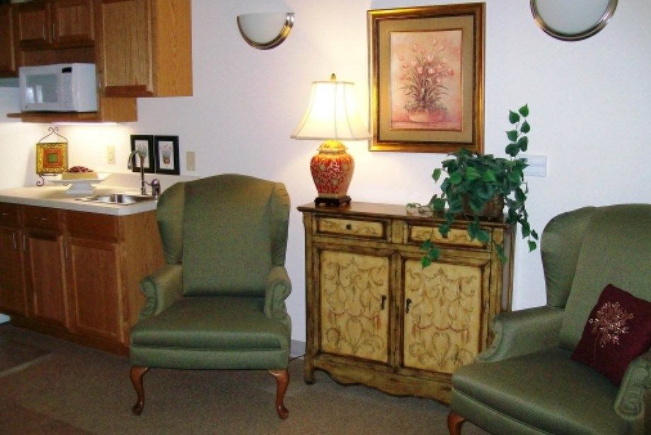 A cozy living room setup featuring green armchairs, a decorative cabinet with a lamp on top, and artwork on the wall.