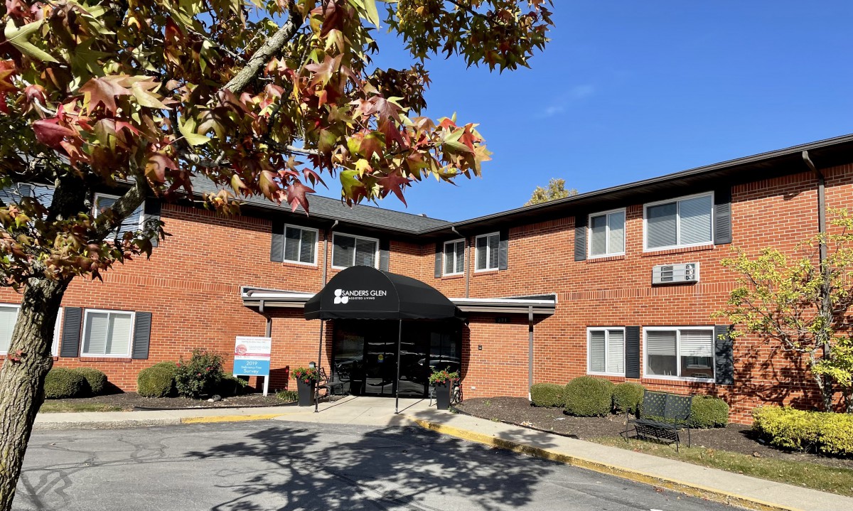 A brick building entrance with an awning, surrounded by autumn trees and a clear blue sky.