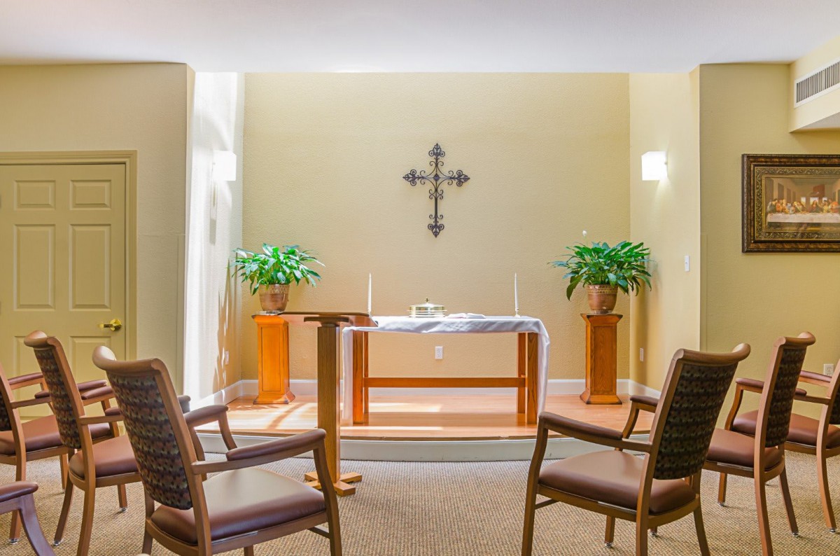 A small chapel with an altar, featuring a cross on the wall, flanked by two plants and surrounded by chairs.
