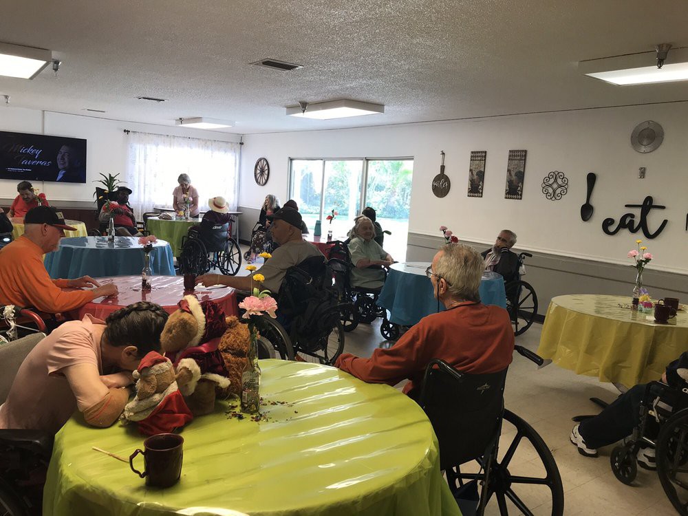 A group of elderly individuals in wheelchairs gathered in a nursing home, with tables adorned with flowers.