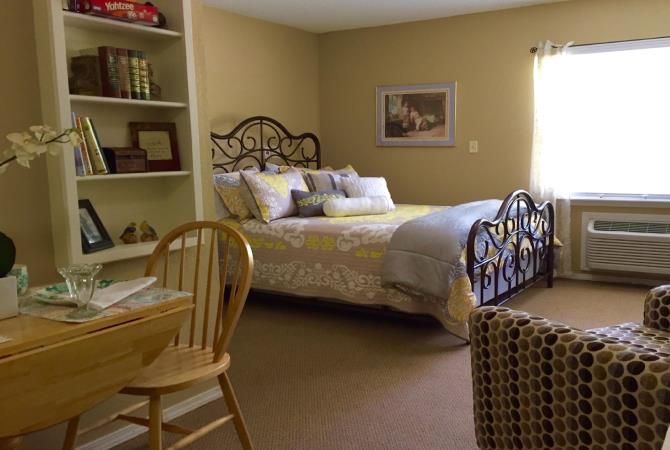 A cozy bedroom with a well-decorated bed, bookshelves, and a wooden chair by the window.