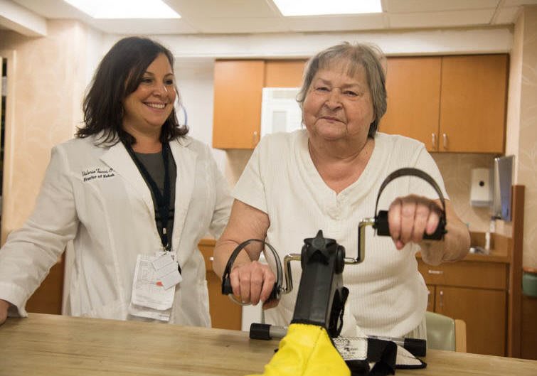An elderly woman engages in a therapy session with a healthcare worker assisting her on exercise equipment.