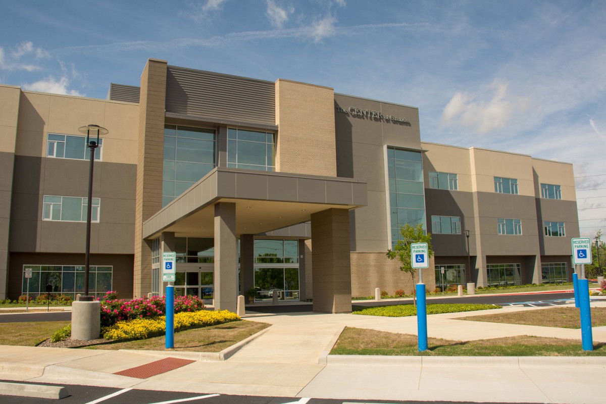 A modern building with large glass windows and a landscaped entrance, featuring designated parking spaces under a clear blue sky.