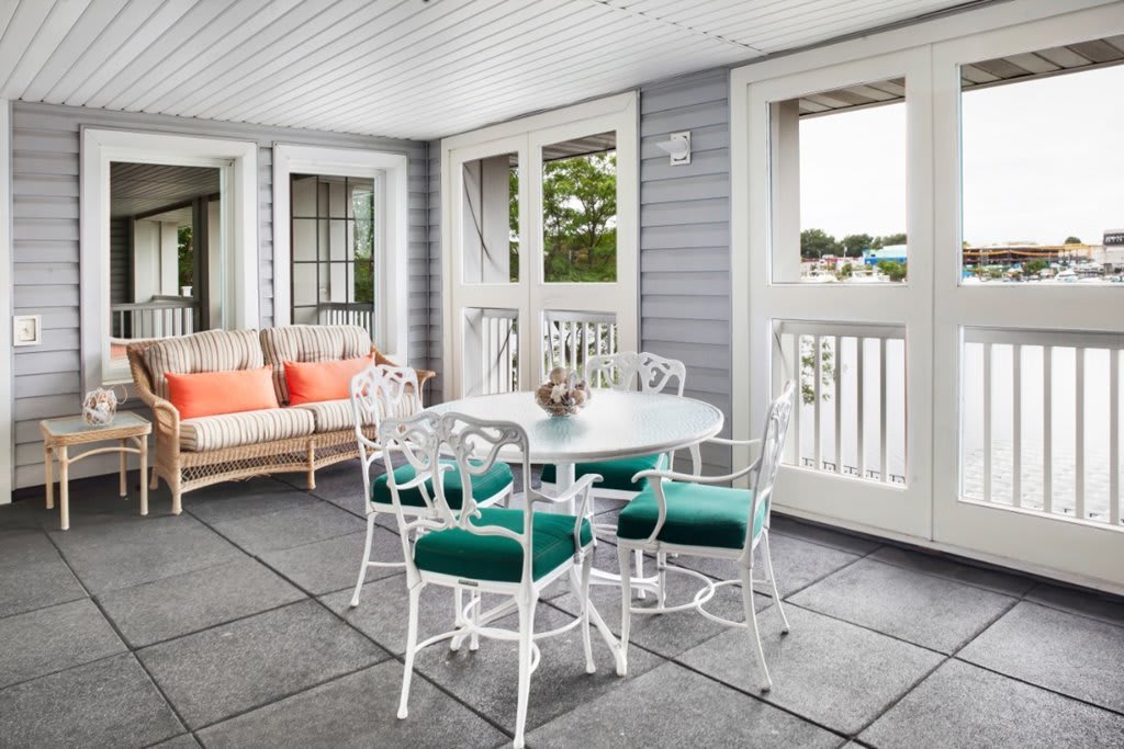 A cozy patio with a white dining set and comfortable sofa, featuring colorful cushions and ample natural light from large windows.