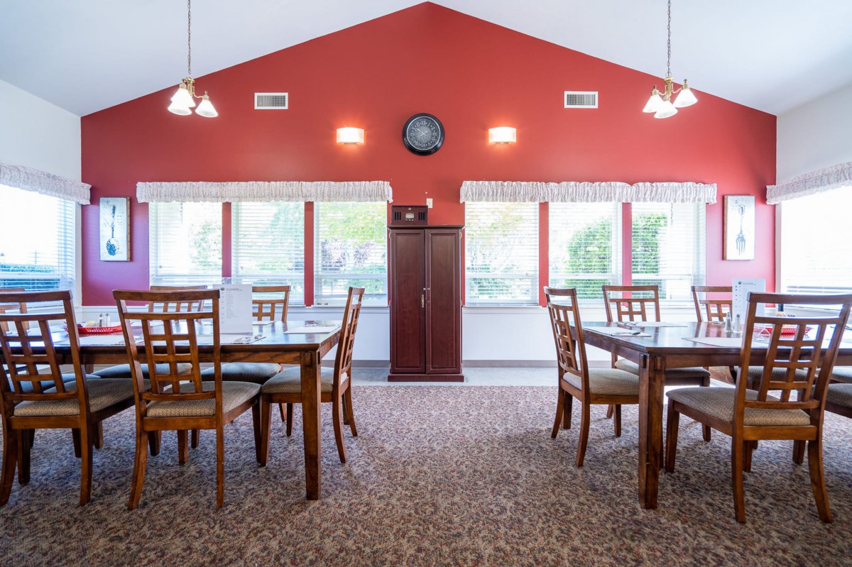 A well-lit dining area with red walls, featuring wooden tables and chairs arranged on a carpeted floor.