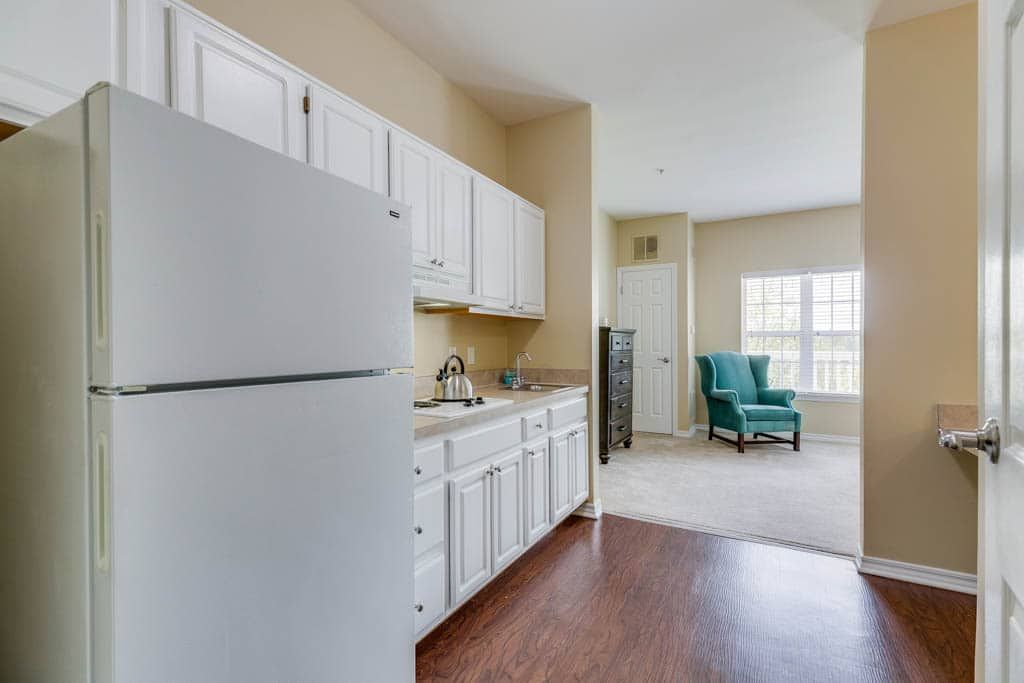 A cozy kitchen and living area with white cabinets, a refrigerator, hardwood floor, and a comfortable chair by the window.